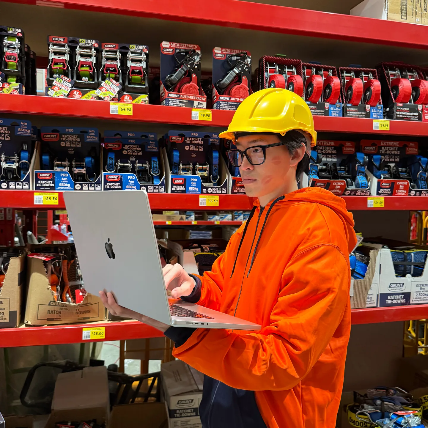A developer in a hard hat and hi-vis hoodie working on a laptop in a hardware store aisle.